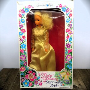 May include: A vintage doll in its original box. The doll is wearing a gold dress with lace sleeves and has blonde hair. The box is white with floral designs and the text "Flower Princess Starry-eyed Bride". The doll is approximately 29 cm tall.