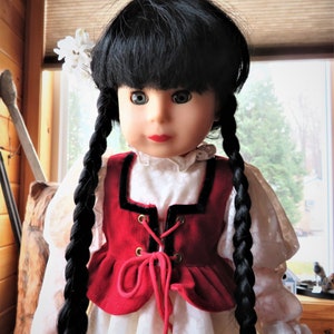 May include: A doll with long black braids, wearing a white blouse with a red velvet vest with black trim and a lace-up front. The doll has a white flower in her hair and is looking at the camera.