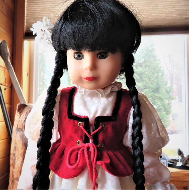 May include: A doll with long black braids, wearing a white blouse with a red velvet vest with black trim and a lace-up front. The doll has a white flower in her hair and is looking at the camera.