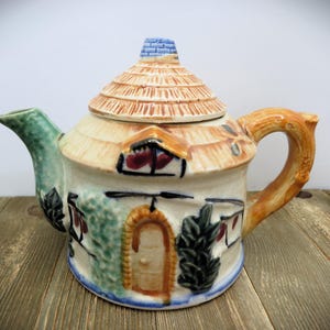 May include: A ceramic teapot shaped like a cottage, featuring a thatched roof lid and a handle resembling a tree branch. The teapot is painted with a light blue, green, brown, and orange color scheme, with details like windows and a door.