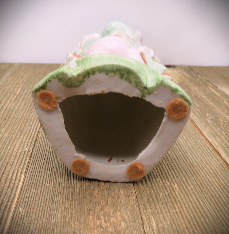 May include: The bottom of a porcelain figurine with a green, pink, and white design. The figurine has four felt pads on the bottom.