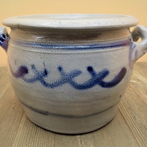 May include: A large, gray stoneware crock with two handles and a blue and white painted design. The crock is sitting on a wooden surface.