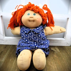 May include: A soft-bodied doll with bright orange yarn hair styled in pigtails with white ribbons. The doll has green eyes, a small dimple on the left cheek, and wears a blue dress featuring a white circular pattern with a collar.
