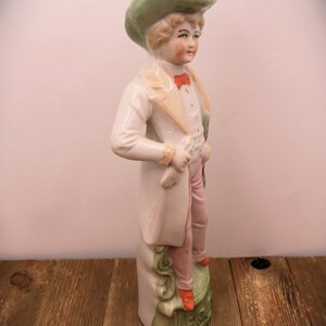 May include: A porcelain figurine of a person wearing a green hat, white coat, and pink pants. The figurine is standing on a green and white base.