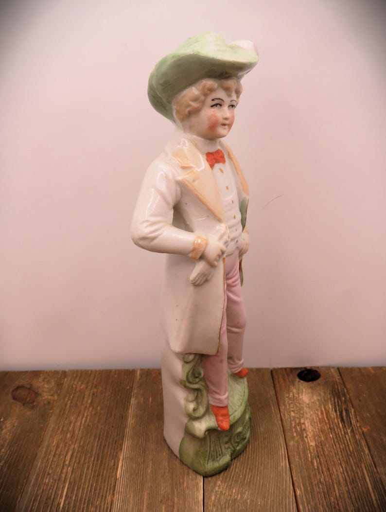 May include: A porcelain figurine of a person wearing a green hat, white coat, and pink pants. The figurine is standing on a green and white base.