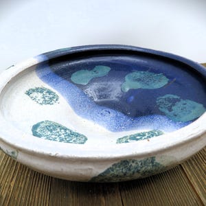 May include: A handcrafted ceramic bowl with a wide, shallow design. The interior features a dark blue center, surrounded by lighter blue and white sections, with teal-colored accents. The exterior is a creamy white with teal speckles. The bowl sits on a wooden surface.