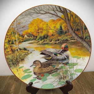 May include: Decorative plate featuring a serene pond scene with two ducks, surrounded by lily pads and reeds. The background showcases a vibrant autumn landscape with a house and trees. The plate has a gold rim.