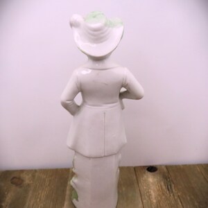 May include: A white porcelain figurine of a person wearing a green and white hat and a long coat. The figurine is standing on a wooden surface.