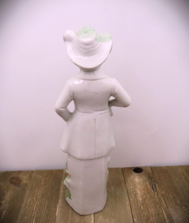 May include: A white porcelain figurine of a person wearing a green and white hat and a long coat. The figurine is standing on a wooden surface.