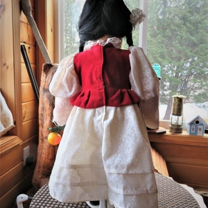 May include: A porcelain doll wearing a white dress with a red vest and a white flower in her hair. The doll is standing on a brown wicker surface.