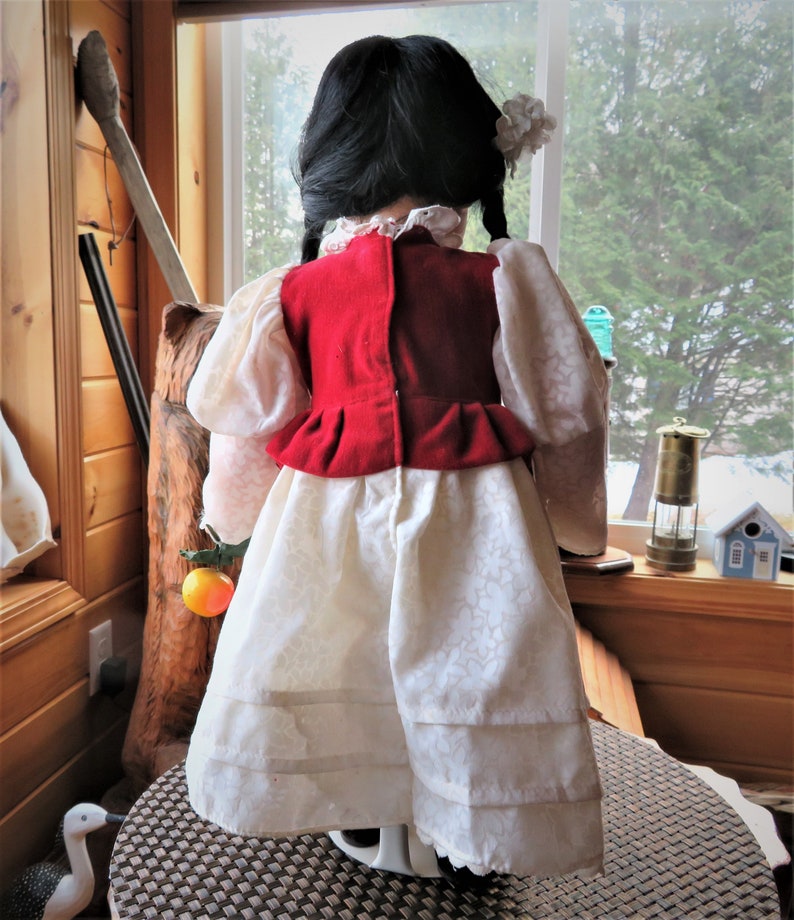 May include: A porcelain doll wearing a white dress with a red vest and a white flower in her hair. The doll is standing on a brown wicker surface.