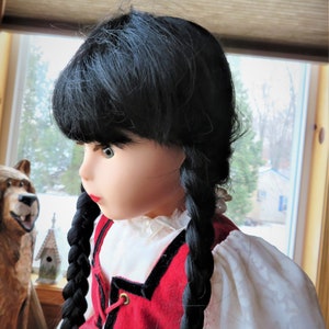 May include: A doll with long black braids wearing a red and black dress with a white blouse. The doll has green eyes and a neutral expression.