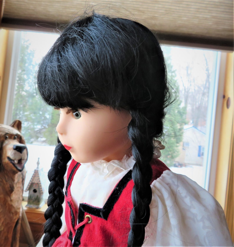 May include: A doll with long black braids wearing a red and black dress with a white blouse. The doll has green eyes and a neutral expression.