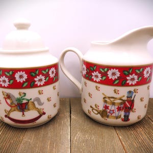 May include: A white ceramic sugar bowl with a lid and a creamer pitcher. Both pieces feature a red band with white floral designs and illustrations of a rocking horse and a toy soldier. The set is on a wooden surface.