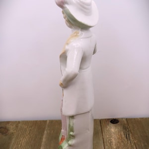 May include: A porcelain figurine of a person wearing a white hat with green and pink accents. The figurine is standing on a base with green and pink floral details. The figurine is painted in white, green, pink, and yellow.