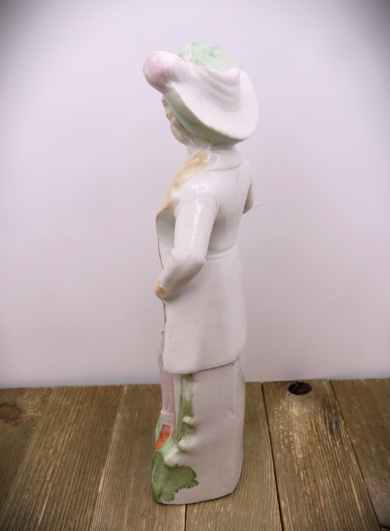 May include: A porcelain figurine of a person wearing a white hat with green and pink accents. The figurine is standing on a base with green and pink floral details. The figurine is painted in white, green, pink, and yellow.