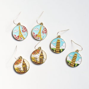 May include: Set of six gold-tone earrings with enamel charms featuring iconic landmarks: the Eiffel Tower, Big Ben, and the Leaning Tower of Pisa. Each charm is surrounded by a blue, white, or yellow background.