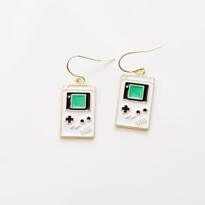 May include: A pair of gold-tone earrings featuring a white enamel design of a retro handheld video game console. The console has a green screen, black accents, and a directional pad. The earrings are a fun, nostalgic accessory.