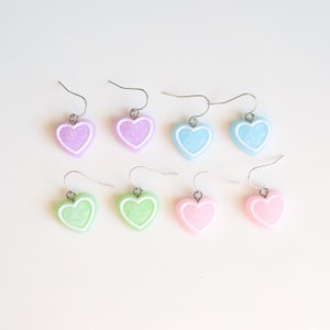 May include: Six pairs of heart-shaped earrings in pastel colors. The earrings are made of a textured material and have a white outline. The colors are light purple, light blue, light green, and light pink.