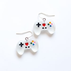 May include: A pair of white, translucent earrings shaped like video game controllers. Each controller has a red heart, black plus sign, and colorful buttons.