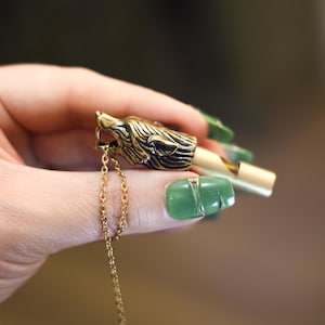 以下が含まれることがあります： 金色の金属製のホイッスルで、オオカミの頭のデザインです。ホイッスルには小さなチェーンが付いています。オオカミの頭は毛皮と顔の特徴が詳細に描かれています。緑色のマニキュアを塗った手でホイッスルを持っています。