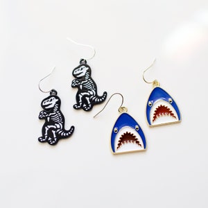 Pode incluir: Um par de brincos pendentes com um esqueleto de dinossauro preto e um tubarão azul com a boca aberta branca e vermelha. Os brincos têm ganchos dourados.