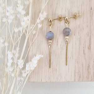 Könnte beinhalten: Ein Paar goldene Ohrringe mit einem grauen Edelstein und einer goldenen Kette. Die Ohrringe liegen auf einem hellen Holzuntergrund.