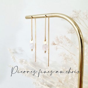 Op de afbeelding: Drie gouden kettingoorbellen met een kleine witte steen en een gouden schijfcharme die aan elke ketting hangt. De oorbellen worden getoond op een gouden standaard tegen een witte achtergrond.