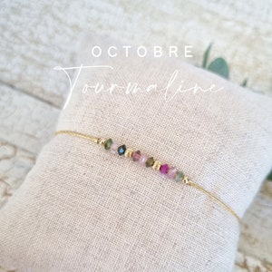 Op de afbeelding: Een gouden armband met een delicate ketting en een rij kleine, kleurrijke edelstenen. De armband wordt getoond op een wit kussen en de tekst "OKTOBER Toermalijn" is zichtbaar boven de armband.