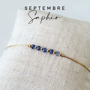 Peut inclure: Un bracelet délicat en chaîne dorée avec une rangée de petites perles de saphir bleu. Le bracelet est sur un fond en tissu blanc. Le texte "SEPTEMBER Saphir" est visible au-dessus du bracelet.