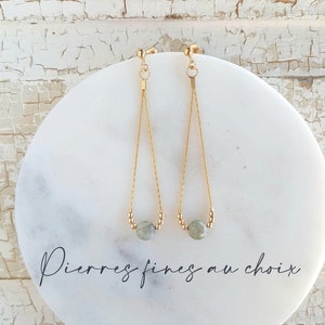 Könnte beinhalten: Ein Paar goldene Ohrhänger mit je einem grauen Stein. Die Ohrringe liegen auf einer weißen Marmorfläche mit dem Text "Pierres fines au choix".