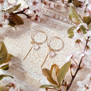 Könnte beinhalten: Ein Paar goldene Creolen mit zarten Ketten und rosa Stein-Akzenten. Die Ohrringe werden vor einem weißen Hintergrund mit einem Blumenarrangement präsentiert.