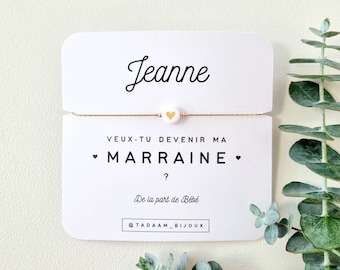 May include: A delicate gold-colored bracelet with a small white heart charm with a gold heart in the center. The bracelet is displayed on a white card with the name "Jeanne" and the text "VEUX-TU DEVENIR MA MARRAINE ?" printed in black.