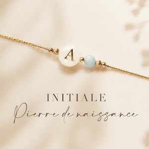 May include: A delicate gold-toned chain bracelet featuring a white circular bead with the letter A in gold, a small light blue spherical bead, and gold-toned accents. The text INITIALE Pierre de naissance is printed below the bracelet.
