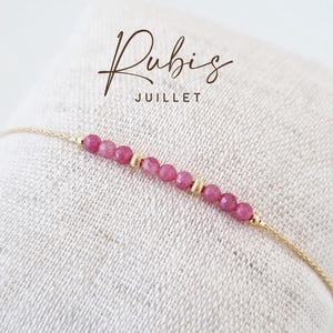 Peut inclure: Un bracelet délicat en chaîne dorée composé d'une rangée de petites perles de rubis roses facettées, entrecoupées de deux minuscules perles intercalaires dorées texturées. Le texte Rubis JUILLET est inscrit en brun au-dessus du bijou.