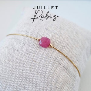 Peut inclure: Un bracelet délicat en chaîne dorée avec une seule perle de pierre précieuse rose. Le bracelet est sur un fond en tissu blanc. Le texte "JUILLET Rubis" est visible au-dessus du bracelet.