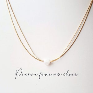 May include: A delicate gold chain necklace with a single white gemstone pendant. The necklace is layered with two chains, creating a simple and elegant look.  "Pierre fine au choix" is written in cursive below the necklace.