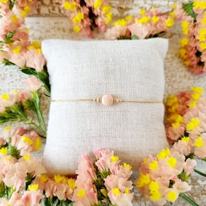 Peut inclure: Un bracelet délicat en chaîne dorée avec une seule perle de pierre précieuse rose. Le bracelet est présenté sur un coussin en tissu blanc entouré de fleurs roses et jaunes.