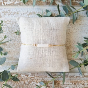 Peut inclure: Un bracelet délicat en chaîne dorée avec une perle rectangulaire blanche et des perles dorées sur un fond en tissu blanc.