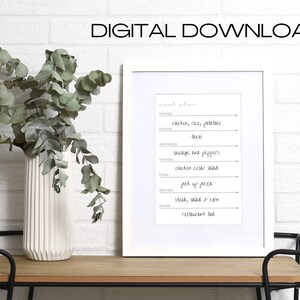 May include: A printable meal planner in a white frame on a wooden table. The planner includes a list of meals for each day of the week, such as "chicken, rice, potatoes" and "steak, salad & corn".