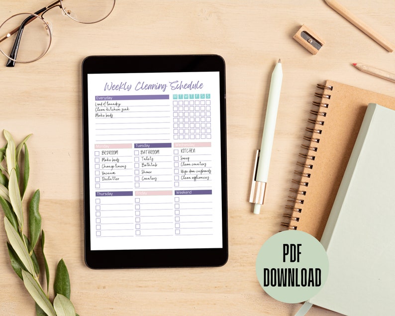 May include: A digital tablet displaying a weekly cleaning schedule with a checklist for everyday, Monday, Tuesday, Thursday, Friday, and Weekend. The schedule includes tasks for the kitchen, bedroom, and bathroom. The tablet is on a wooden surface with a pen, pencil, and a notebook.