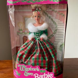 Puede incluir: Una muñeca Barbie con un vestido de cuadros verdes y rojos con cuello y puños de piel blanca. La muñeca está en una caja con las palabras "Winter's Eve Barbie" en la parte delantera.