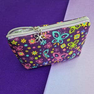 May include: A purple zippered pouch with a butterfly and flower print. The pouch has a silver zipper and a silver charm in the shape of a flower.