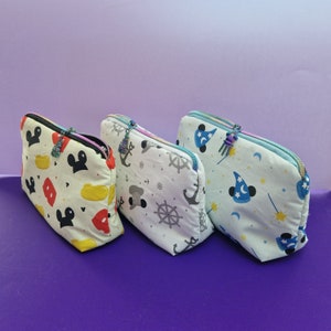 May include: Three white zippered pouches with different Disney character designs. The pouch on the left has Mickey Mouse and Minnie Mouse designs. The middle pouch has a nautical theme with an anchor and a ship's wheel. The pouch on the right has a blue and white design with a wizard hat and a star.