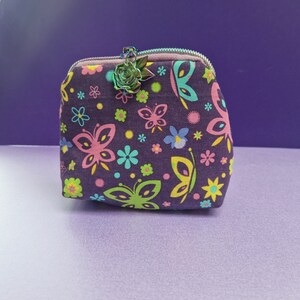 May include: A small, square, zippered pouch with a dark purple background and a colorful butterfly and floral pattern. The zipper pull is a metal rose. The pouch is approximately 4 inches wide and 3 inches tall.