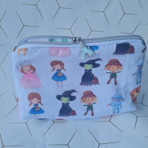 May include: White zippered pouch with a repeating pattern of characters from the Wizard of Oz. The characters include Dorothy, the Wicked Witch of the West, the Scarecrow, the Tin Man, and the Cowardly Lion. The zipper pull is a silver charm shaped like a castle.