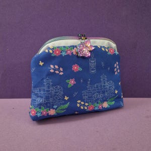 May include: A blue fabric coin purse with a floral and architectural pattern. The purse has a silver zipper and a decorative rose-shaped charm. The design includes pink and yellow flowers, butterflies, and candle illustrations.