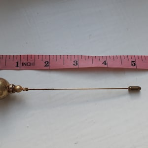 Op de afbeelding: Een gouden speld met een ronde, stervormige, decoratieve bovenkant en een lange, dunne steel. De speld is ongeveer 10 cm lang.