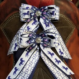 May include: Two navy blue and white horse-print hair bows with pink accents and silver concho buttons. The bows are tied together and displayed on a brown saddle.