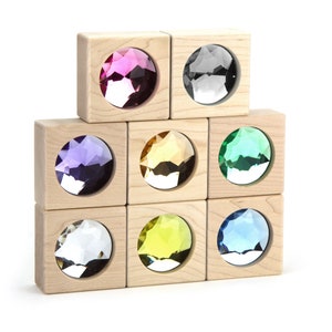 May include: A set of eight light-colored wooden blocks arranged in a stack, each featuring a circular cutout embedded with a faceted, translucent gemstone. The gems display a variety of colors including pink, clear, purple, amber, green, yellow, and blue.